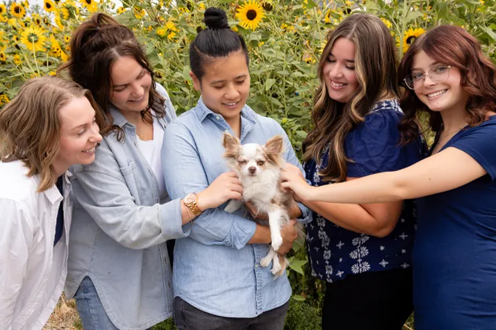 Chiropractor American Fork UT Debbie Pun With Team and Dog Chiropractor, Debbie Pun, holding dog in sunflower field surrounded by team.